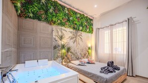 A modern bedroom with a built-in bathtub, a bed with a grey patterned bedspread, and a wall decorated with a green plant wall and a tropical mural. - Asian Oasis with Balneo & Massage (Toulon)
