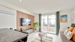 A modern living room with a marble table, a sofa with patterned cushions, a television mounted on a wooden panel wall, and a large window with a view of buildings. - Ô Repos des Artistes - The Gatsby (Toulon)