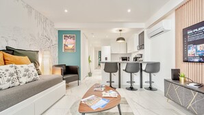 A modern living room with a sofa, coffee table, and bar stools. There is a kitchen area with a microwave and a TV mounted on the wall. - Ô Repos des Artistes - The Gatsby (Toulon)
