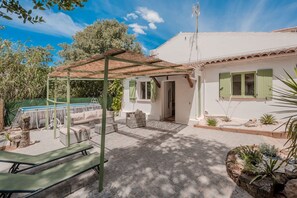 Un bâtiment blanc avec des volets verts, un patio couvert avec des meubles d’extérieur, et une piscine en arrière-plan.