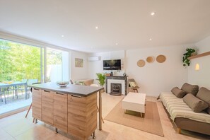 A modern living room with a sofa, a dining area with a table and chairs, a kitchen with a TV and a fireplace, and a large window with a view of greenery. - La Grande Clémente, 2 flats (Toulon)