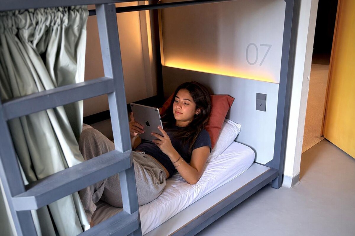 Een stapelbed met iemand die een tablet gebruikt, een zichtbaar kamer nummer '07', en een gordijn dat het raam gedeeltelijk bedekt.