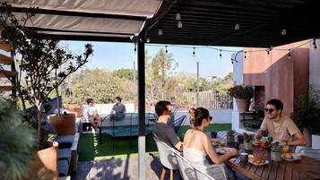 An outdoor patio with wooden tables and chairs, surrounded by potted plants and a swimming pool.