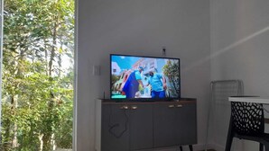 A room with a TV on a cabinet, a dining table, and a view of greenery outside. - Cozy chalet surrounded by nature in a gated community (101) (Parque Rizzo Cotia)