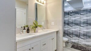 Une salle de bain avec un meuble-lavabo blanc, un grand miroir, un rideau de douche orné de motifs de nuages et une petite plante posée sur le meuble-lavabo.