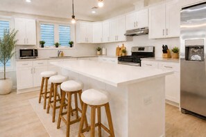 A modern kitchen with white cabinetry, a central island with stools, stainless steel appliances, and potted plants. - Luxury 6BR Rooftop • Free Parking • Near NYC & EWR (Newark)