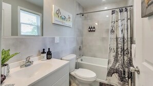 Une salle de bain moderne avec un lavabo, des toilettes et une baignoire blancs. On y trouve un rideau de douche orné d’un motif représentant la Tour Eiffel, ainsi qu’un miroir au-dessus du lavabo.