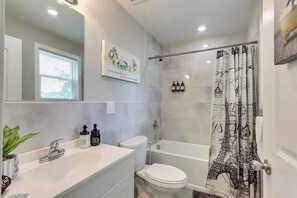 Ein modernes Badezimmer mit Waschbecken, Toilette und Badewanne in Weiß. Ein Duschvorhang mit Eiffelturm-Motiv und ein Spiegel über dem Waschbecken.