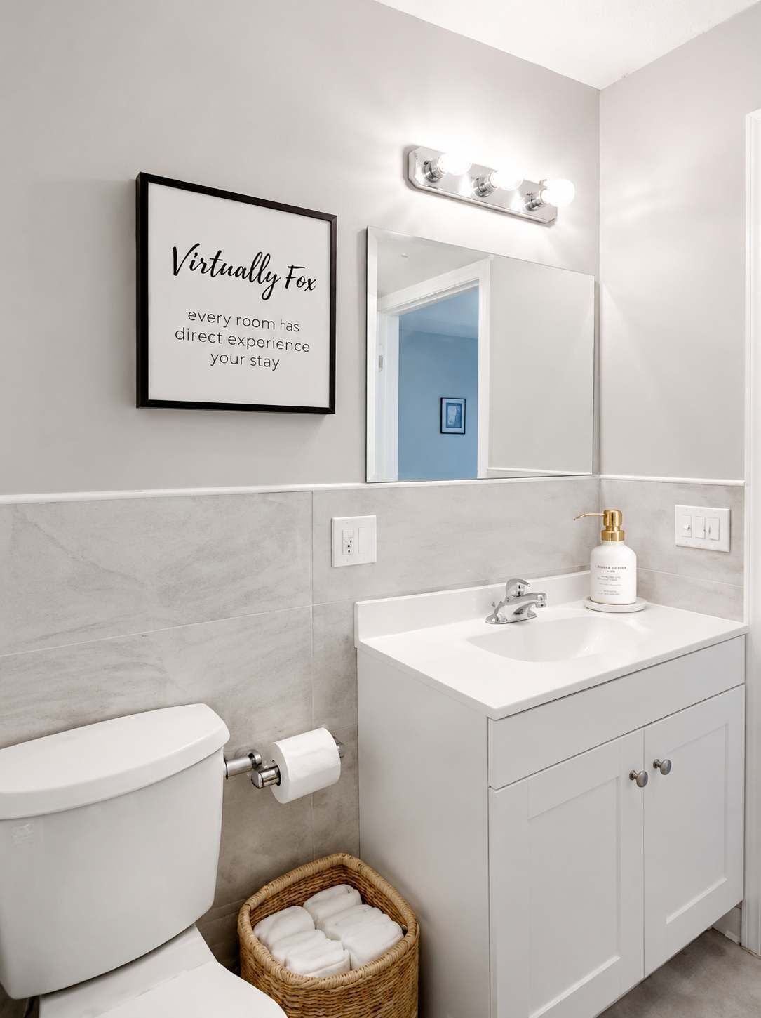 A modern bathroom with a toilet, sink, and a mirror with lights. There's a framed quote on the wall and a wicker basket with towels.