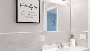 A modern bathroom with a toilet, sink, and a mirror with lights. There's a framed quote on the wall and a wicker basket with towels.