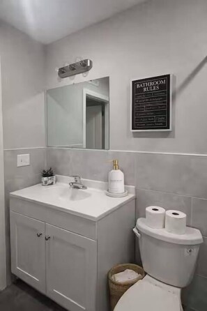 Un baño con lavamanos, espejo y inodoro blancos. Hay un cartel en la pared que dice 'Bathroom Rules'.