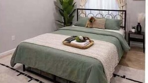 Een slaapkamer met een bed, gordijnen, een plant en een dienblad met spullen op het bed.