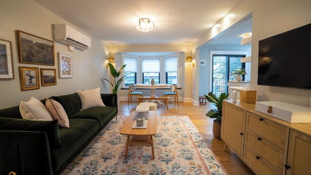 A living room with a green sofa, a wooden coffee table, and a TV on a wooden cabinet. There is a dining area with a table and chairs, and a large window providing natural light. - Prime Location on Main Street (Stroudsburg)