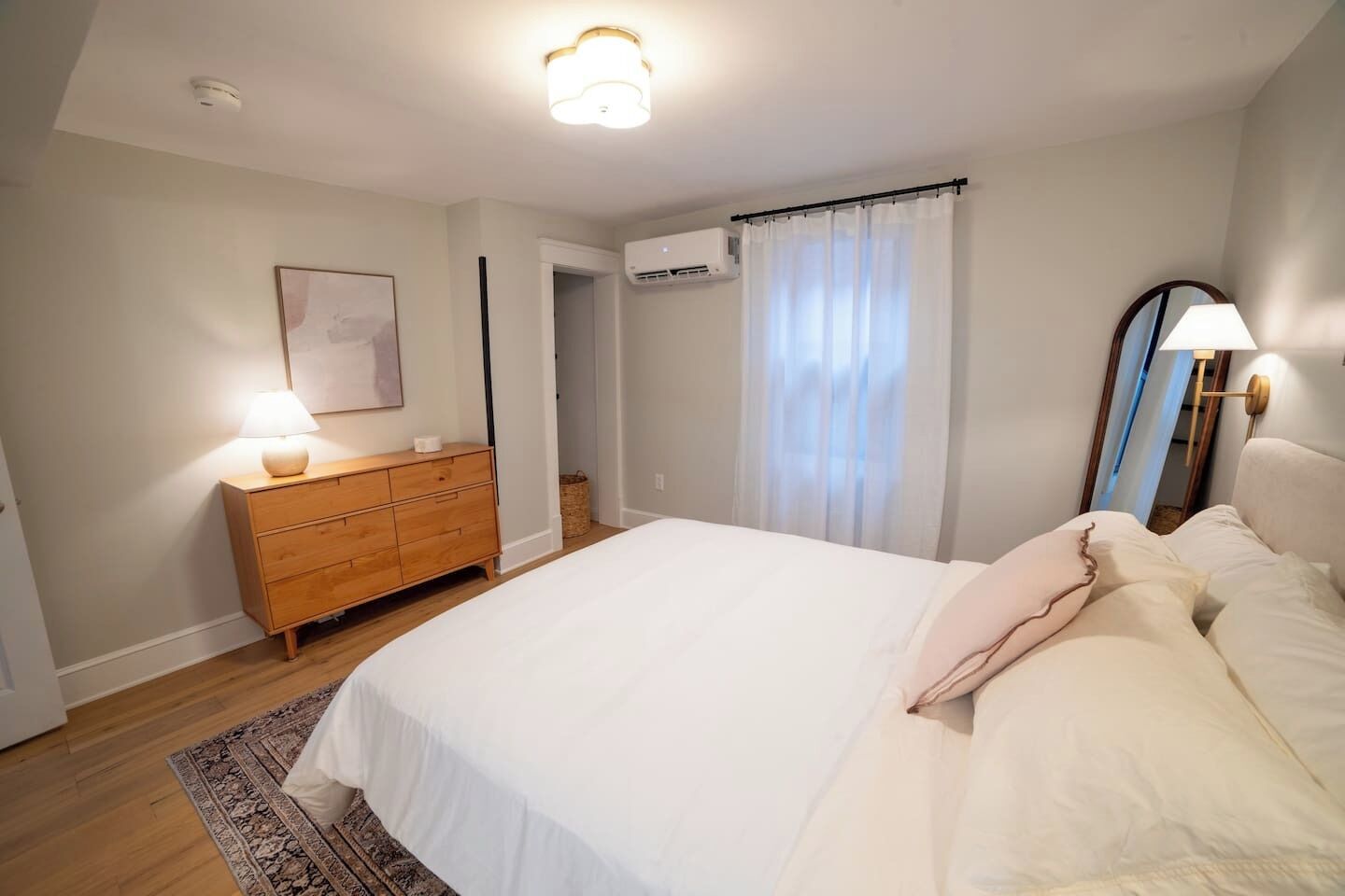 Une chambre à coucher comprenant un lit, une commode, une lampe, un appareil de climatisation et une fenêtre avec des rideaux.