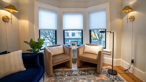 Ein Eckzimmer mit drei großen Fenstern, einem blauen Sofa, zwei beigen Sesseln, einem kleinen Tisch und einem gemusterten Teppich.