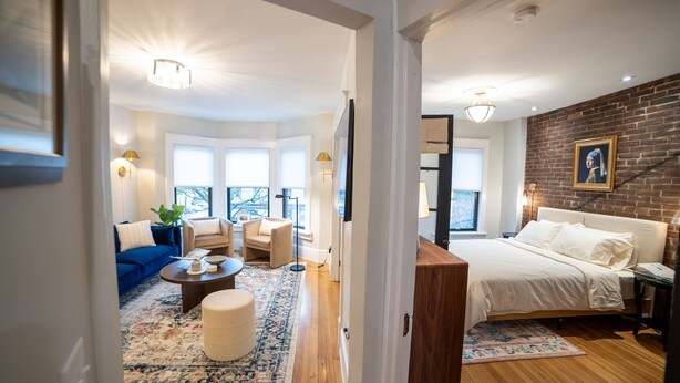 Ein modernes Hotelzimmer mit einem großen Bett, einer Sitzecke mit Sofa und Sesseln, einem Couchtisch und Blick auf die Stadt durch große Fenster.