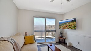 Ein Wohnzimmer mit einer Couch, einer Bank, einem Fernseher und einer Schiebetür, die auf einen Balkon führt.
