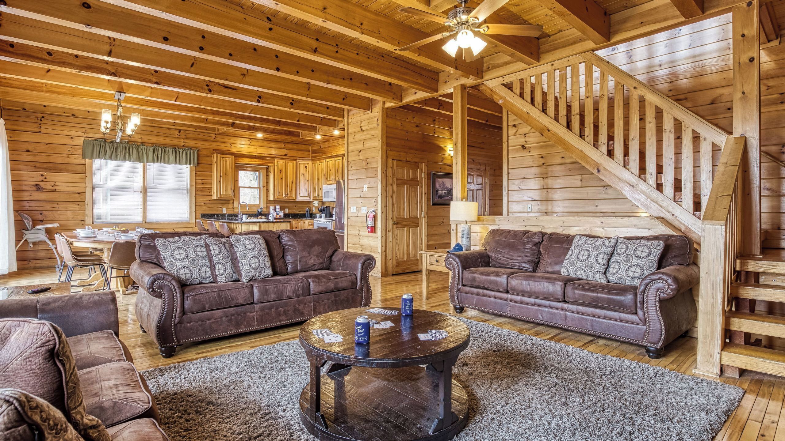 A log cabin interior with two leather sofas, a wooden dining table, a kitchen area, and a staircase.