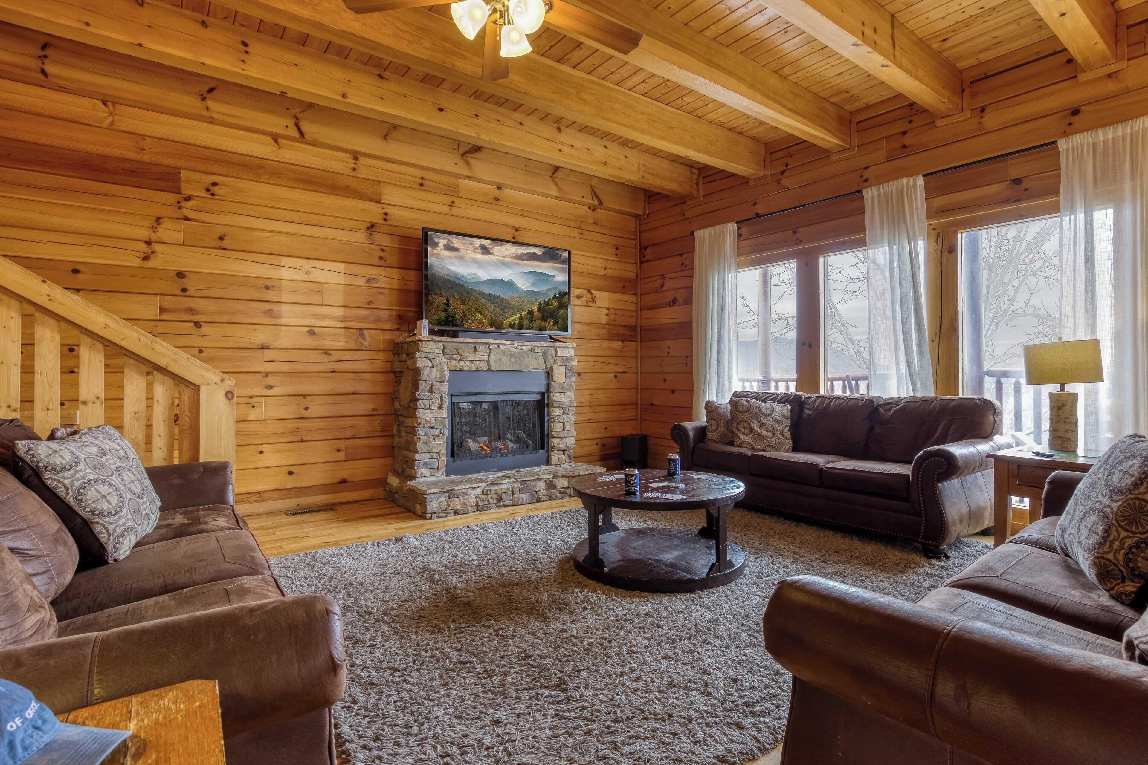 A cozy log cabin interior with a stone fireplace, leather sofas, and a large TV on the wall.
