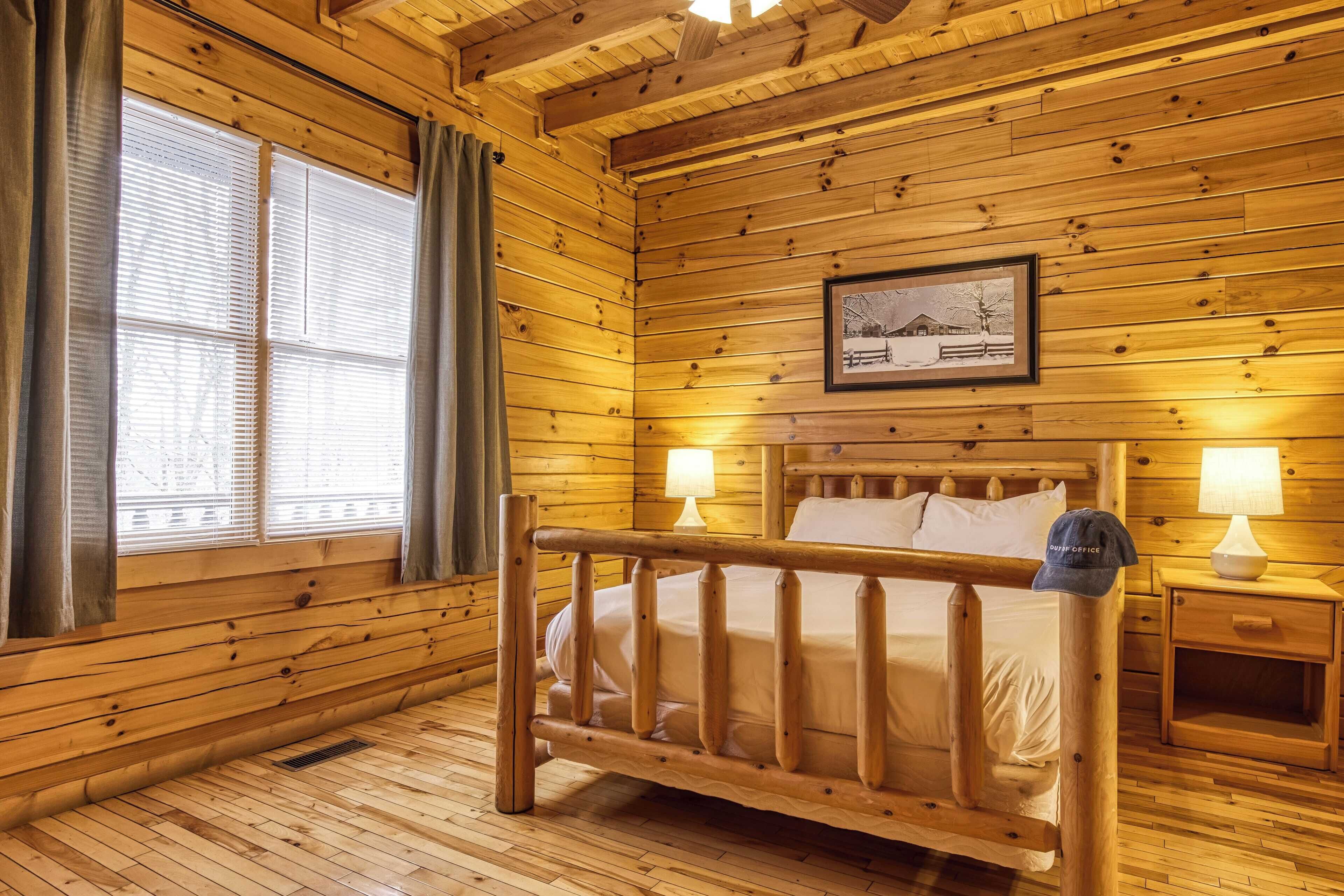 A wooden cabin room with a bed, two bedside lamps, a framed picture on the wall, and a window with curtains.