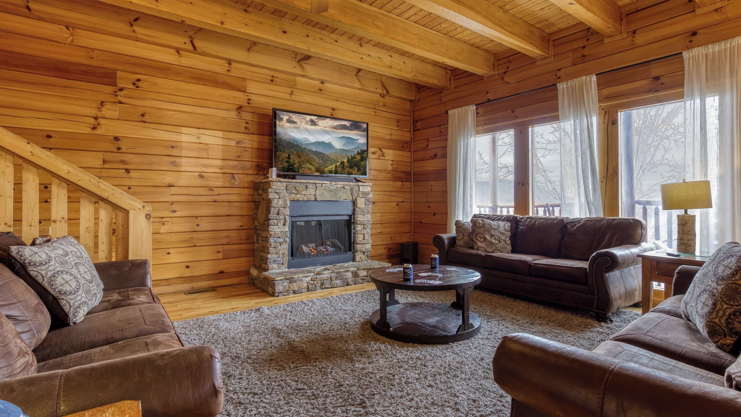 L’intérieur chaleureux d’un chalet avec une cheminée en pierre, des murs en bois, un canapé en cuir et une grande fenêtre.