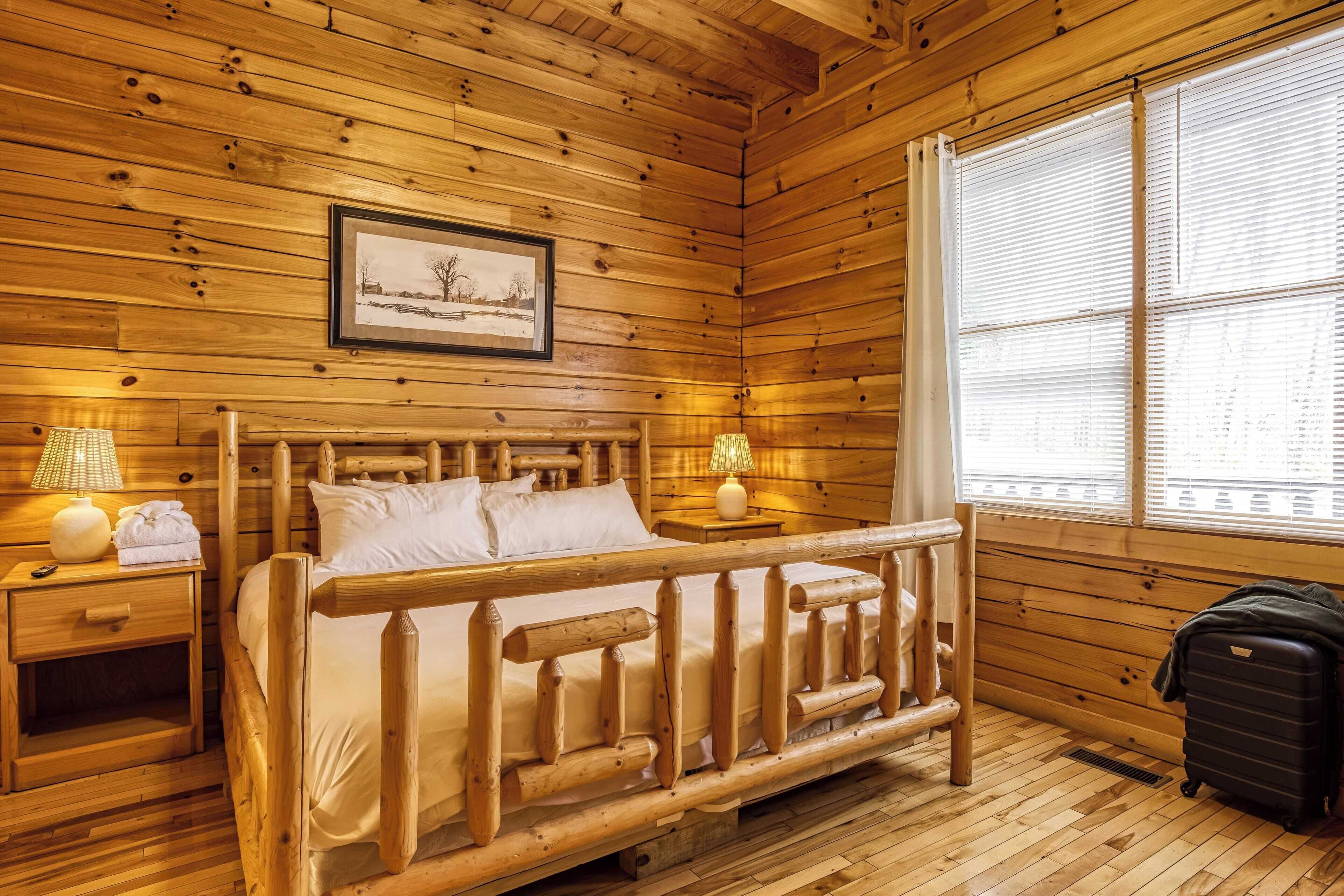A wooden cabin room with a bed, two bedside tables, a window with blinds, and a framed picture on the wall.