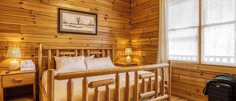 A wooden cabin room with a bed, two bedside tables, a window with blinds, and a framed picture on the wall.
