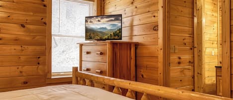 Una habitación de cabaña de madera con una cama, un televisor sobre un aparador de madera y una ventana con persianas.