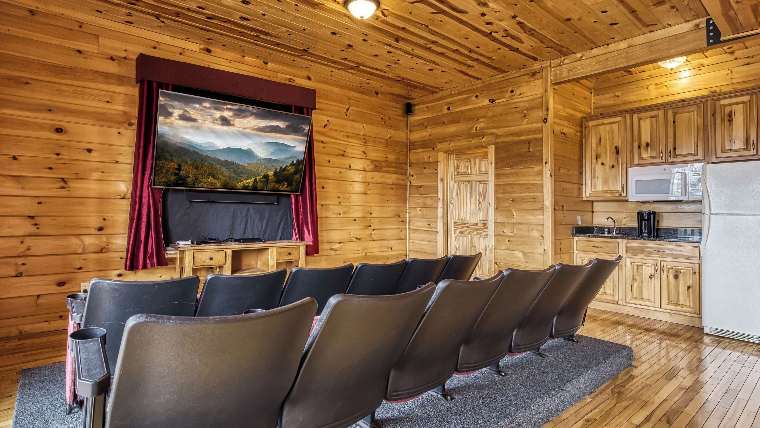 A cozy cabin interior with a movie screen, seating area, kitchen, and wooden walls.