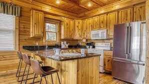 A wooden cabin-style kitchen with a central island, bar stools, a stainless steel refrigerator, and wooden cabinets. - Lookout Lodge by Avantstay Sleeps 18 Hot Tub Game Room Movie Theater View (Sevierville)