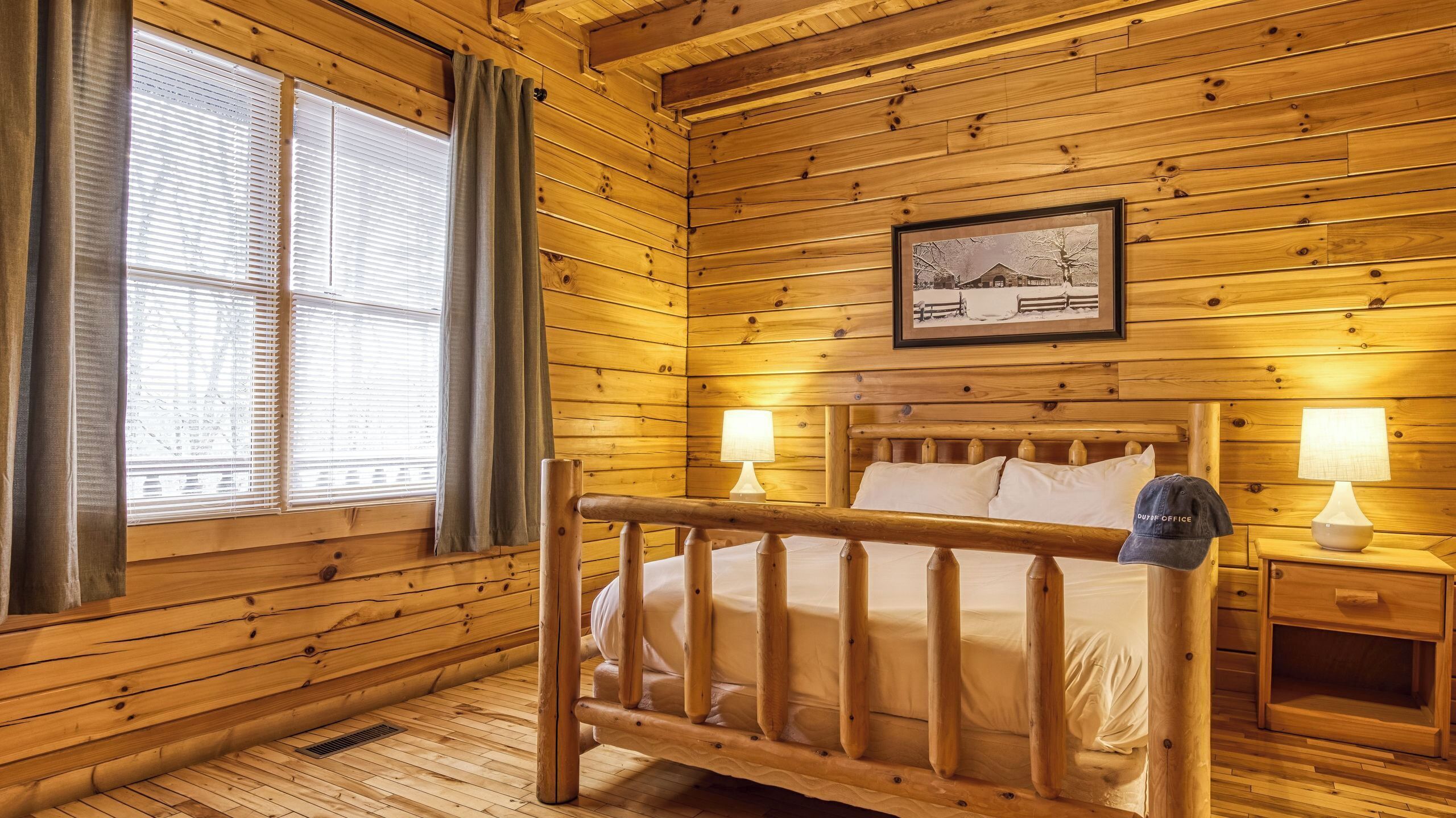 A wooden cabin room with a bed, two bedside lamps, a framed picture on the wall, and a window with curtains.