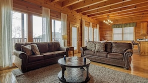 A cozy cabin interior with two leather sofas, a round coffee table, and a kitchen area visible through a doorway. - Lookout Lodge by Avantstay Sleeps 18 Hot Tub Game Room Movie Theater View (Sevierville)