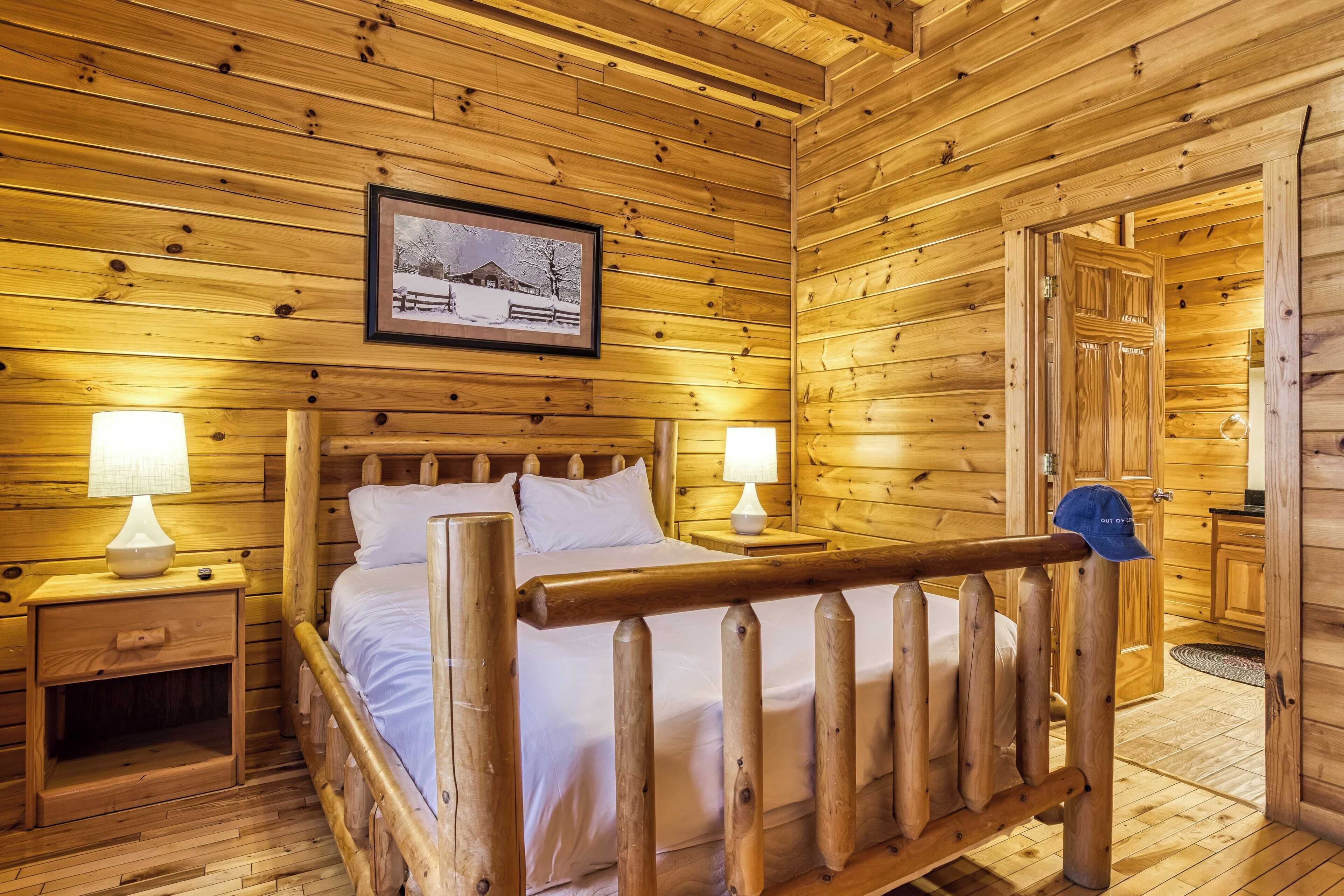 A wooden cabin room with a bed, two bedside tables with lamps, a framed picture on the wall, and a door leading to another room.