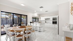 A modern kitchen with white cabinetry, a central island, and a dining area with white chairs and a wooden table. - Water Lane - Swimming Pool - 4 Mins Walk to Bay (Callala Bay)