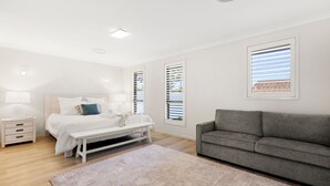A modern bedroom with a bed, bedside tables, a bench, a grey sofa, and large windows with blinds. - Water Lane - Swimming Pool - 4 Mins Walk to Bay (Callala Bay)