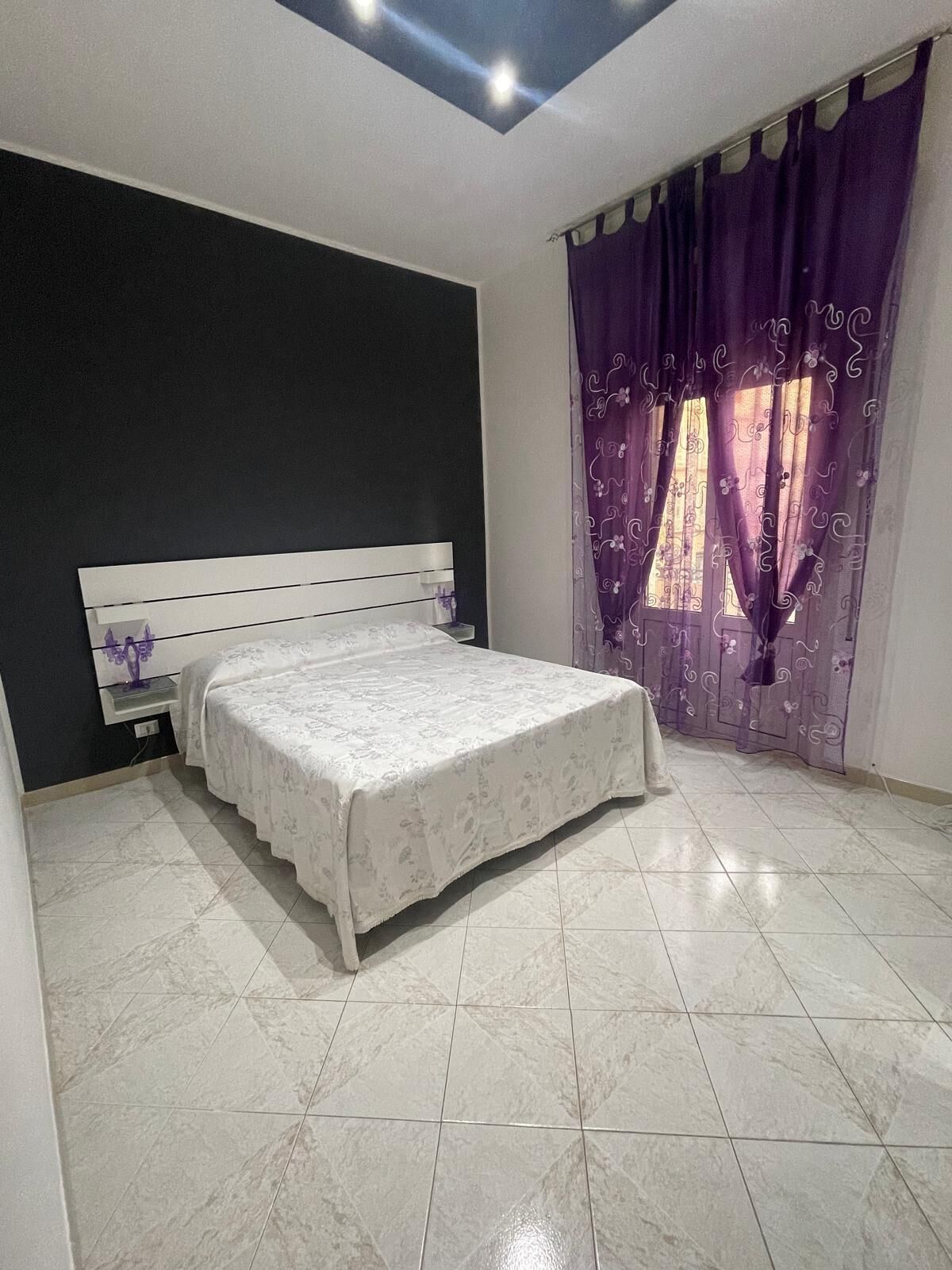 Una camera da letto con un letto, un comodino, una finestra con tende viola e un lucernario.