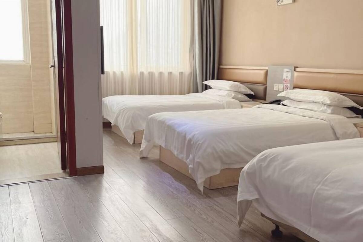 Habitación de hotel con tres camas individuales, suelo de madera, ventana con cortinas traslúcidas y un termostato en la pared.