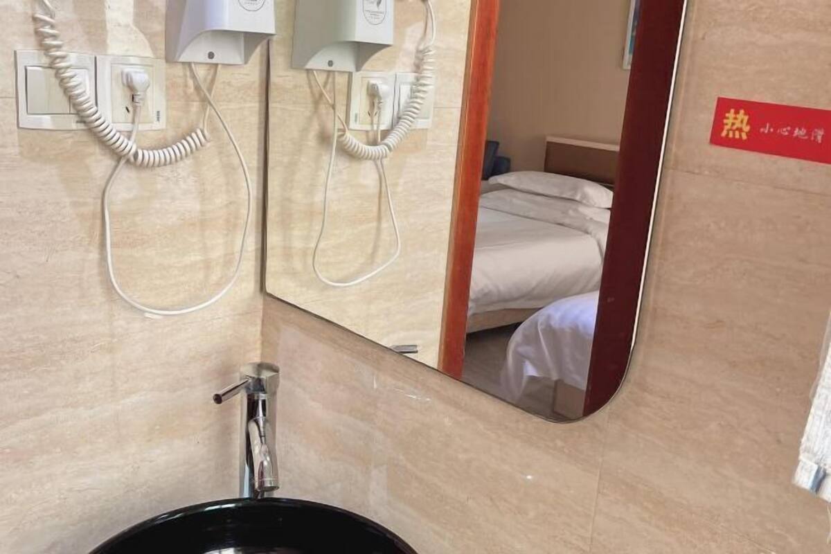 Un baño con un lavamanos, dos secadores de pelo montados en la pared y un espejo que refleja una cama con ropa blanca.
