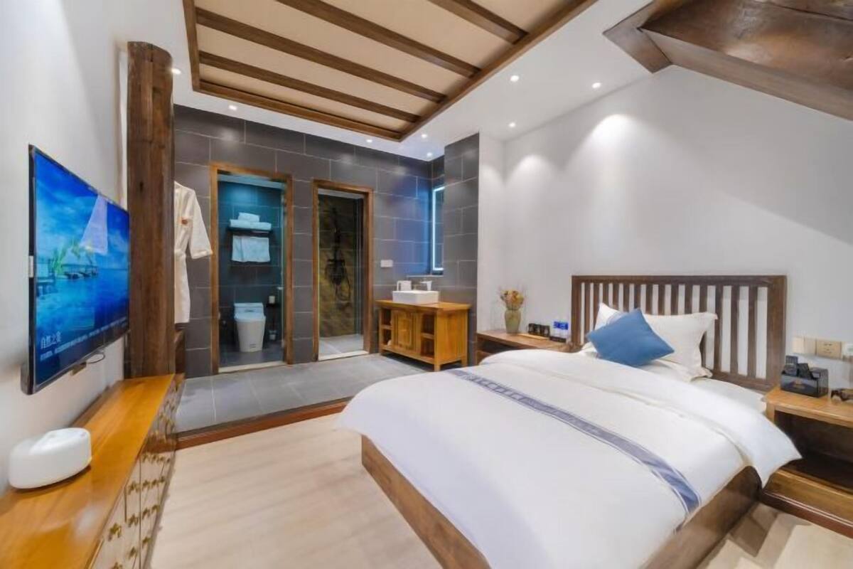 A modern hotel room with a large bed, a wooden headboard, a TV, and a bathroom with a glass door.