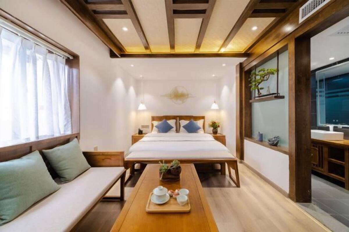 A modern hotel room with a bed, bedside tables, a wooden bench, a tray with tea set, and a window with curtains.