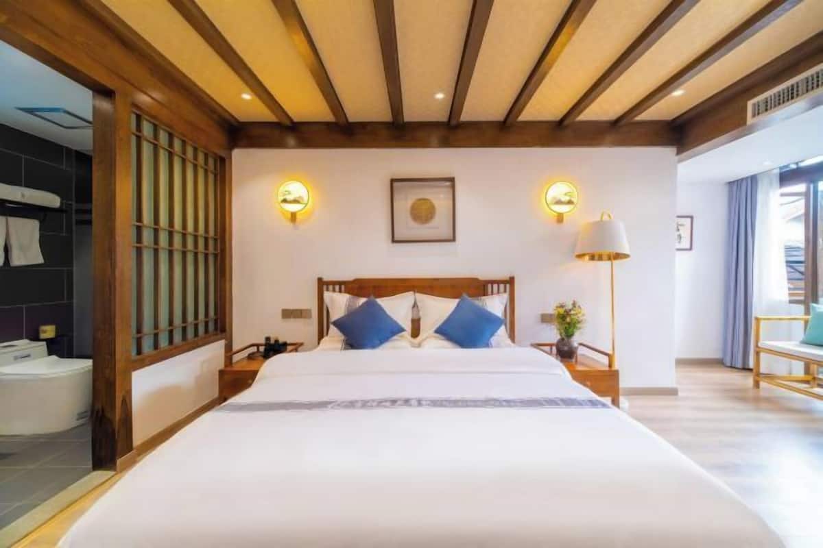 A hotel room with a wooden ceiling, a bed with white bedding and blue pillows, a bedside table with a lamp, and a view of the outside through a window.