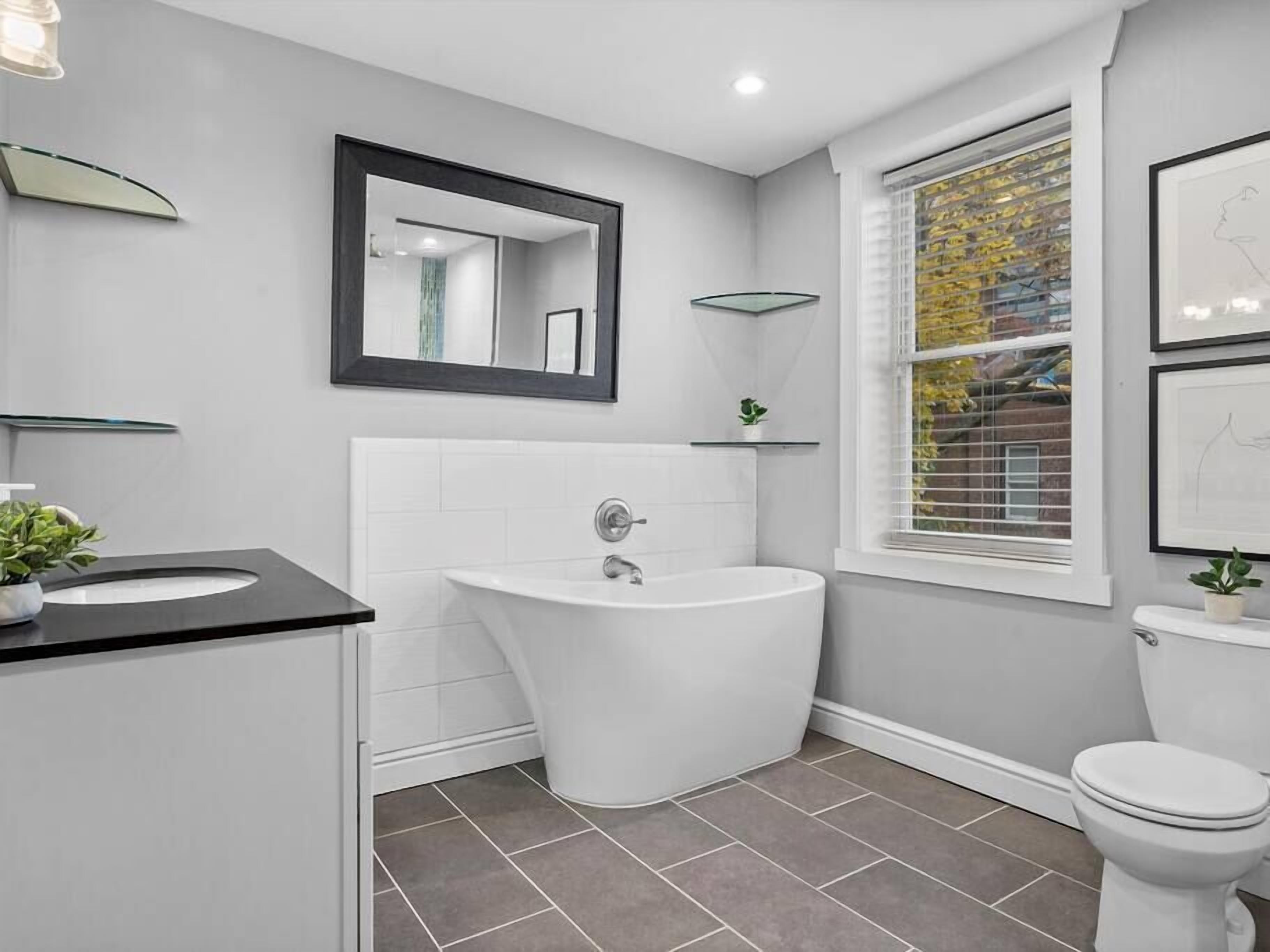 A modern bathroom with a freestanding bathtub, a white sink, and a toilet. There is a window with blinds, a mirror above the sink, and a small shelf with a plant.
