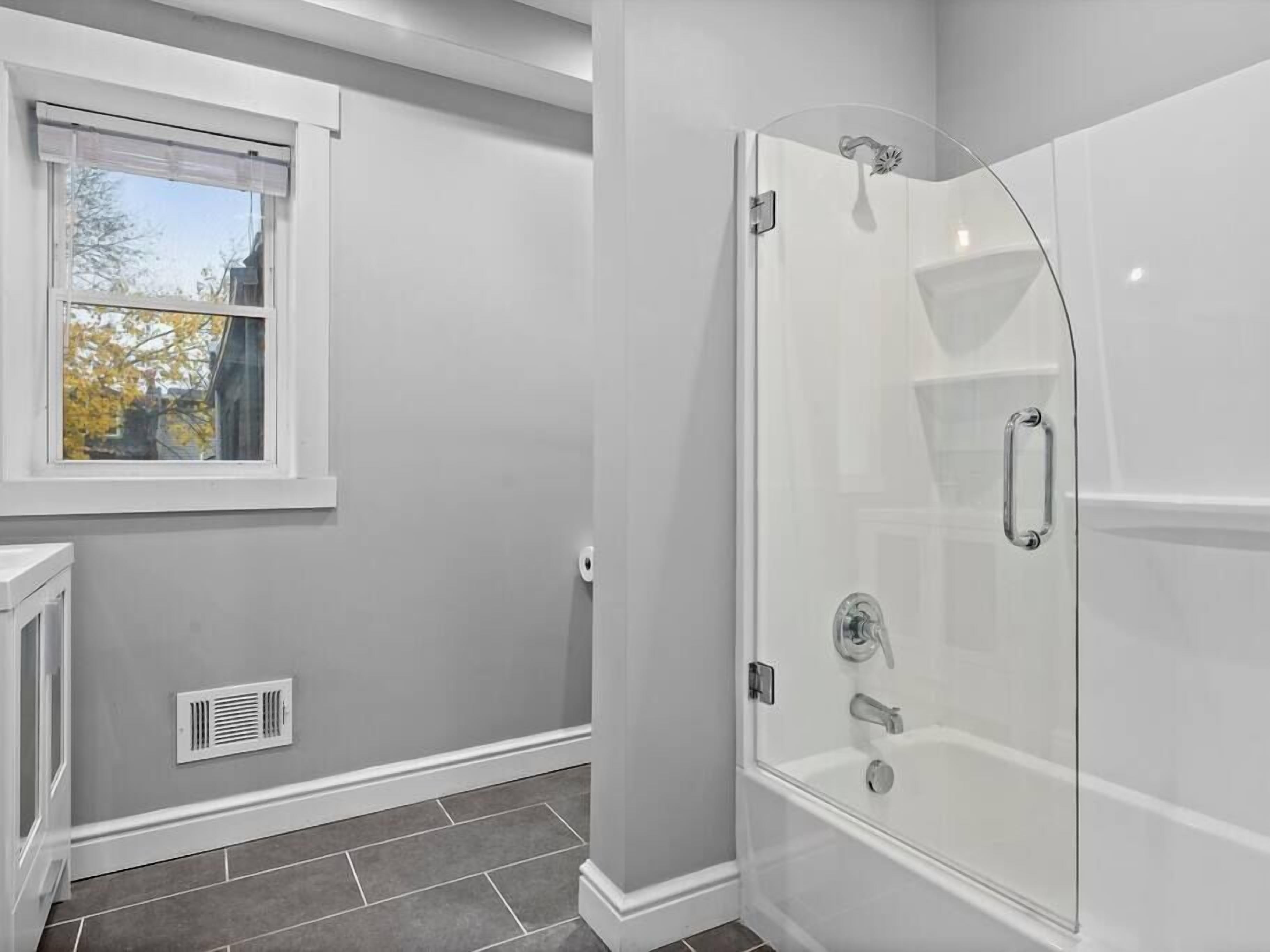 A modern bathroom with a walk-in shower, a window with a view of trees, and a vent on the wall.