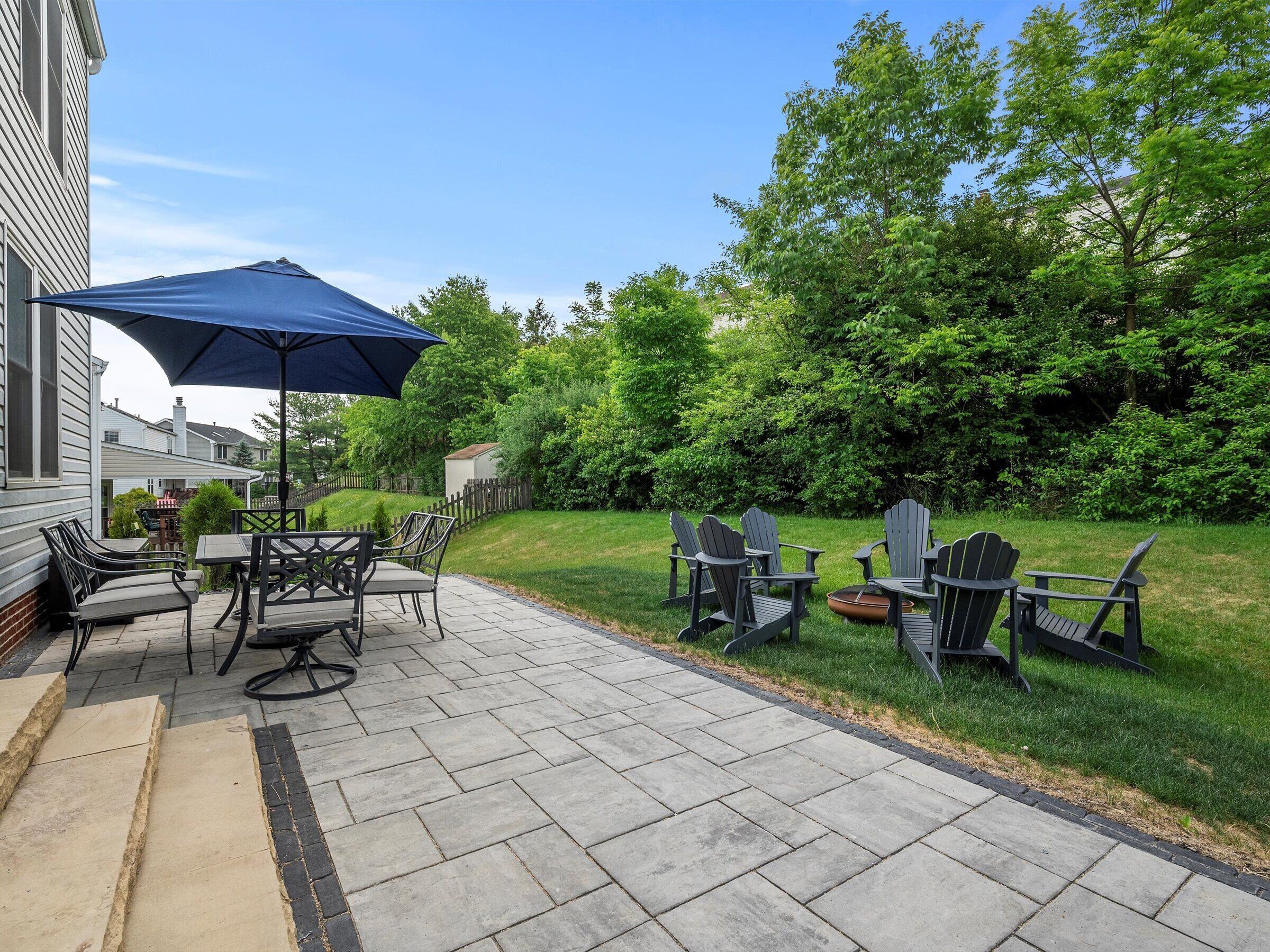 Une terrasse extérieure avec une table et des chaises, un parasol bleu et une pelouse agrémentée de fauteuils Adirondack.