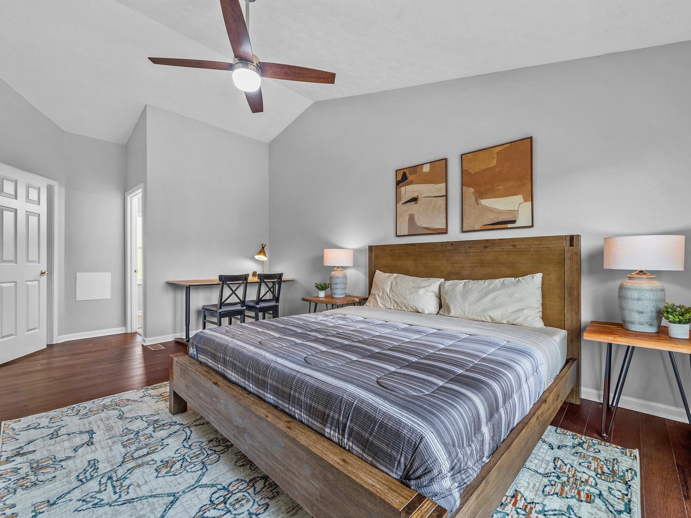A bedroom with a wooden bed, a ceiling fan, two bedside tables with lamps, a desk with chairs, and abstract wall art.