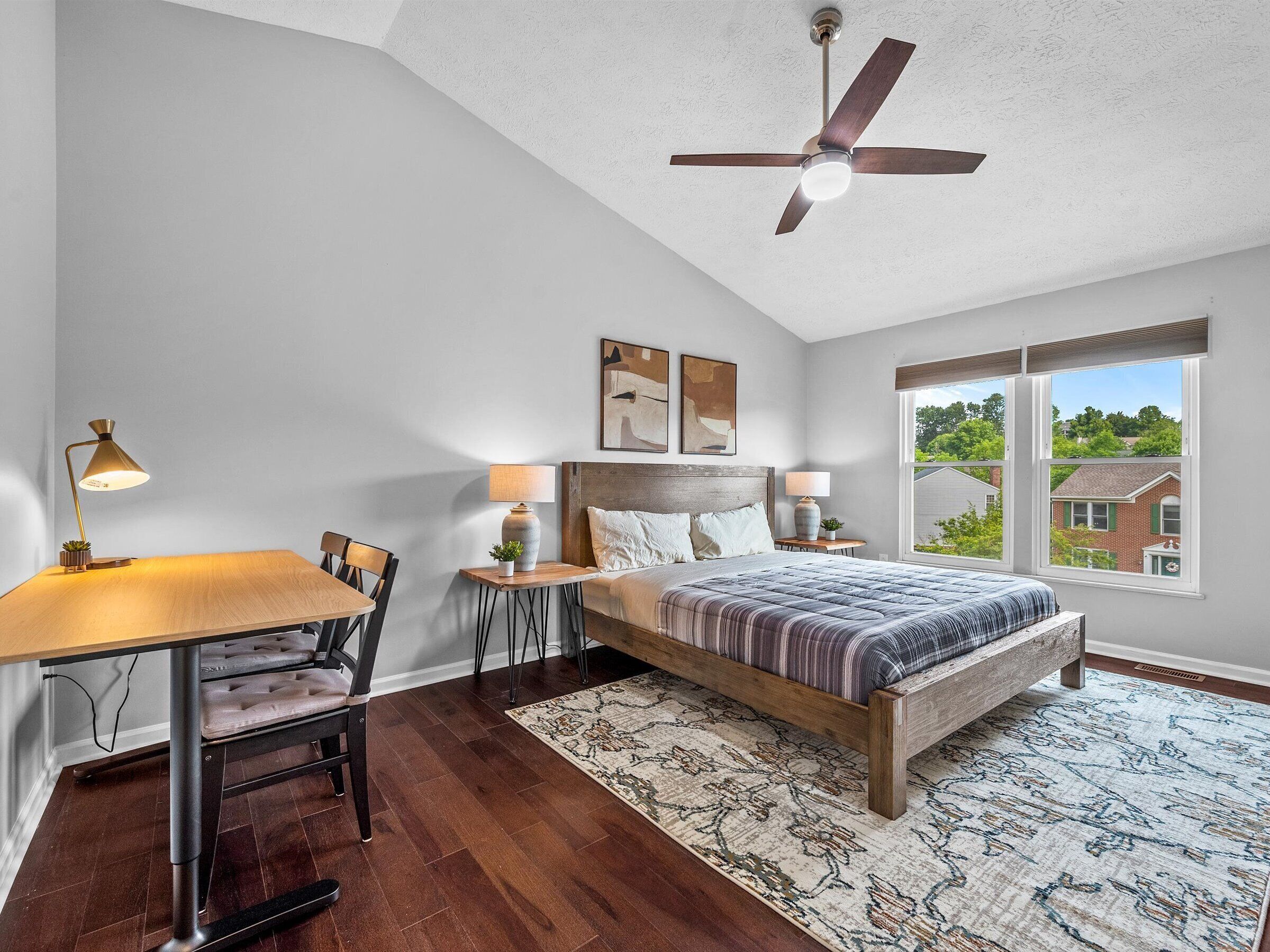 Une chambre à coucher comprenant un lit, un bureau, une chaise, un ventilateur de plafond et une fenêtre donnant sur des maisons.
