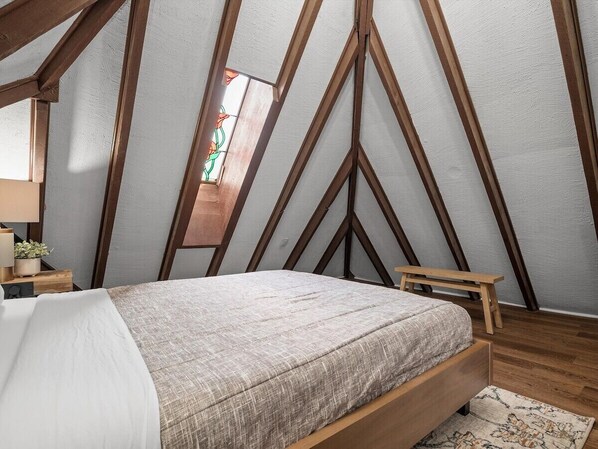 Une chambre avec un plafond en pente, des poutres en bois, un lit recouvert d’une couverture blanche et grise, un petit tabouret en bois et une table de chevet avec une lampe et une plante.