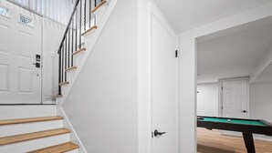 Una escalera con peldaños de madera que conduce a una puerta blanca, una mesa de billar en la habitación contigua y una vista de otra puerta al fondo.