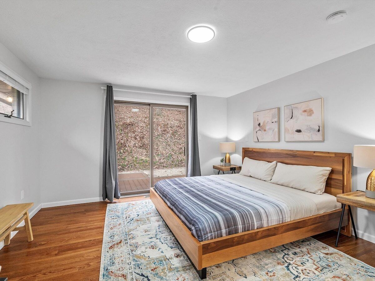 Un dormitorio con una cama de madera, un banco, un ventanal con cortinas y dos lámparas de pared.