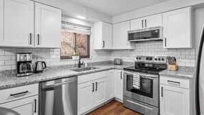Una cocina moderna con gabinetes blancos, electrodomésticos de acero inoxidable, lavavajillas y un ventanal con persianas.